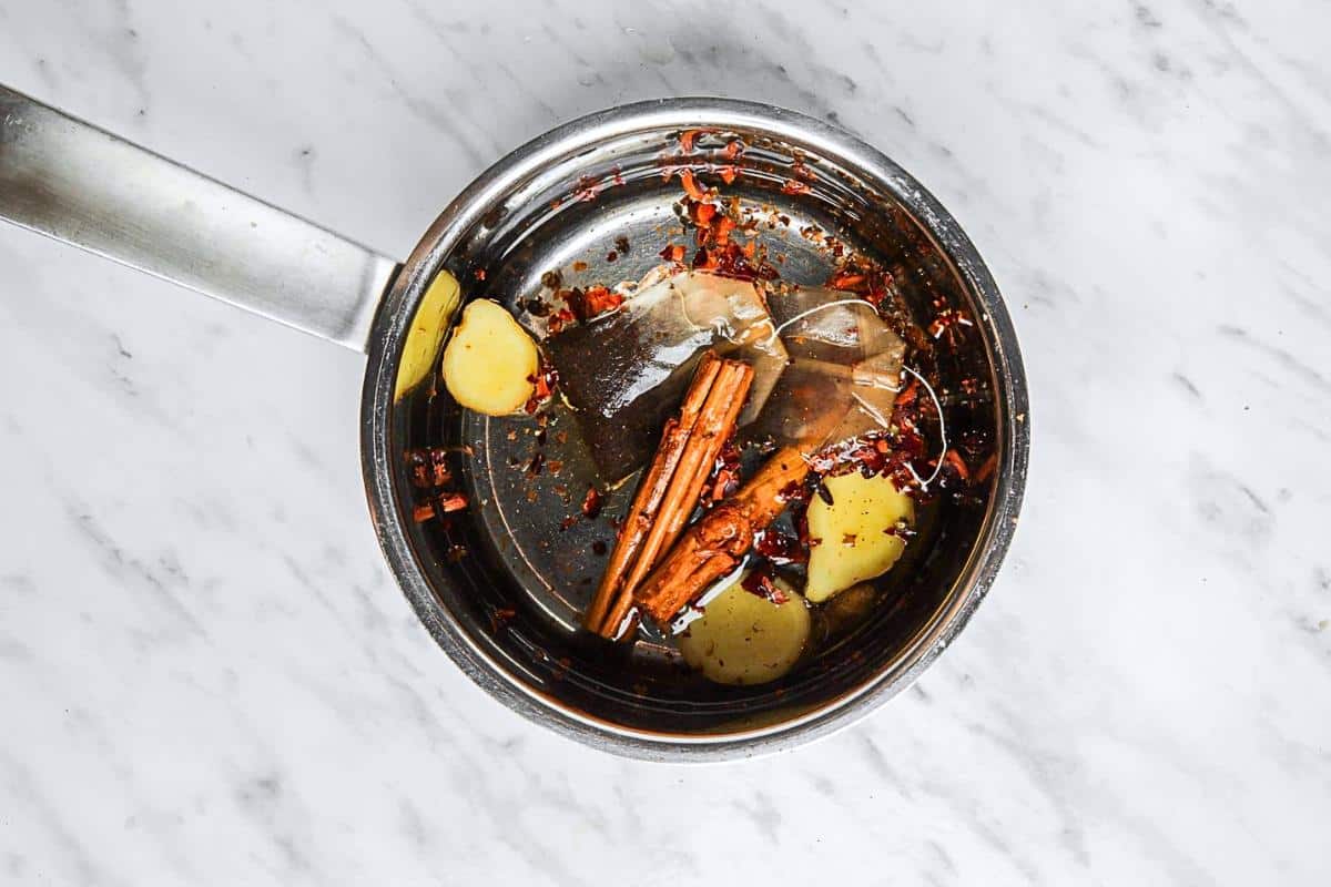 A small saucepan with a metal handle containing water, sliced ginger, cinnamon sticks, and tea bags on a marble surface.