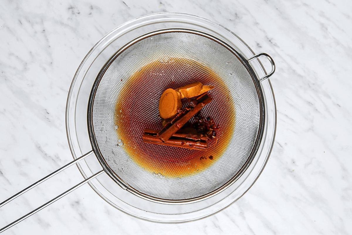 A metal strainer containing cinnamon sticks, cloves, and ginger slices sits over a glass bowl. Dark liquid drains through the strainer, resting on a white marble countertop.