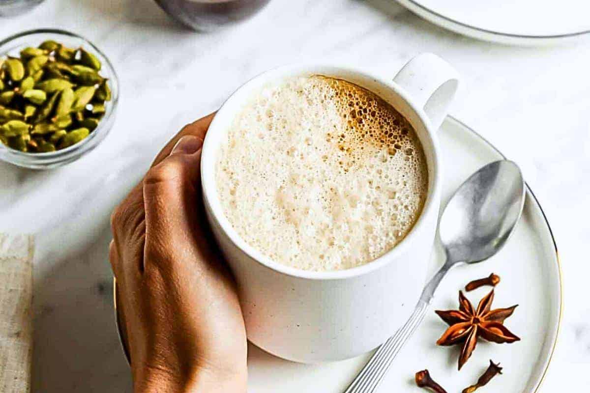 A person's hand holding a white cup filled with frothy chai tea on a white saucer, accompanied by a silver spoon. Star anise and cloves are on the saucer, and a small bowl of cardamom seeds is nearby on the white marble surface.