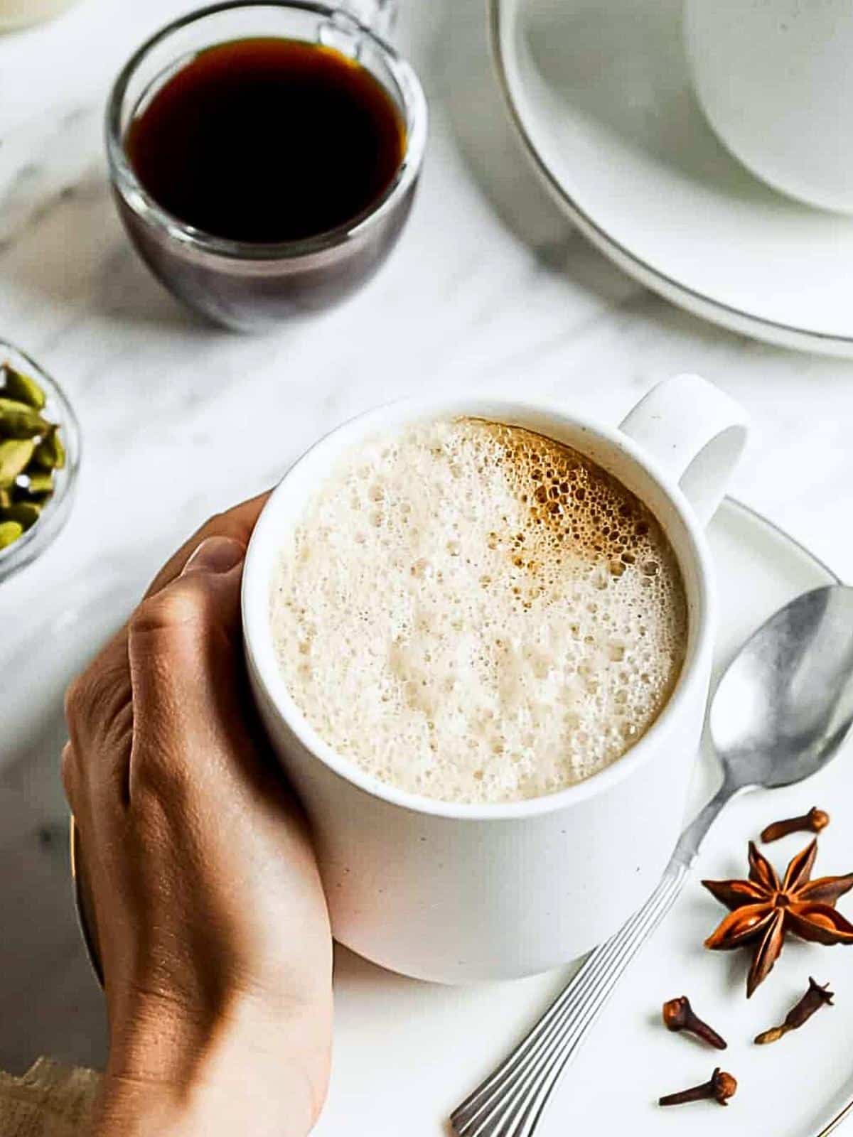 A hand holds a frothy cup of coffee on a white plate with a spoon. Nearby, there's a small bowl of cardamom pods, a glass cup of black coffee, and scattered star anise and cloves on a marble surface.