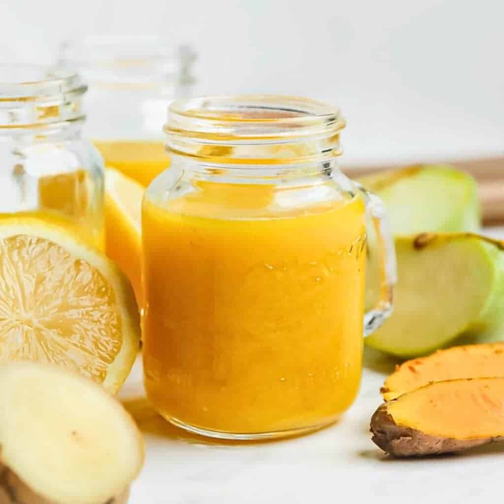 A glass jar filled with vibrant orange juice is surrounded by fresh ingredients: lemon slices, ginger, and a piece of turmeric. Another jar is partially visible in the background. The setup is on a light surface.