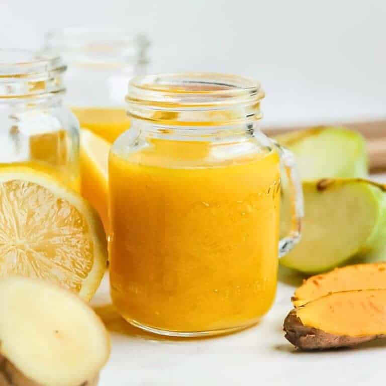 A glass jar filled with vibrant orange juice is surrounded by fresh ingredients: lemon slices, ginger, and a piece of turmeric. Another jar is partially visible in the background. The setup is on a light surface.