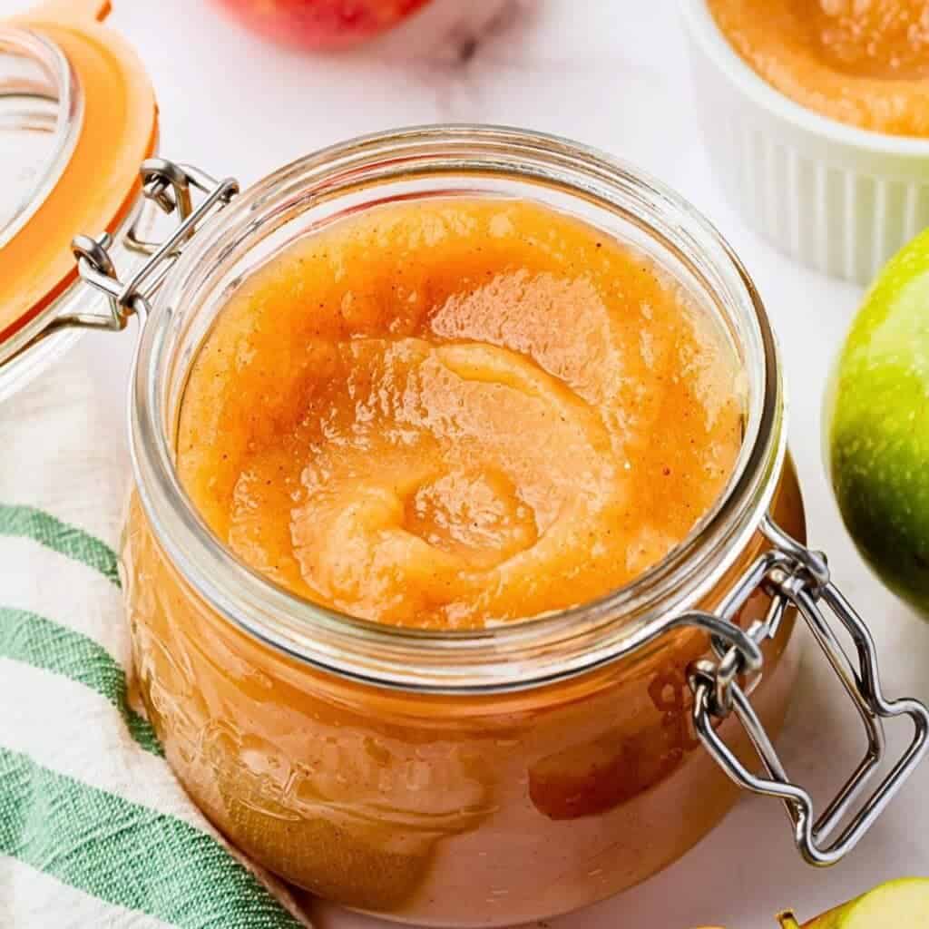 A glass jar filled with smooth, golden applesauce. It's open with a metal clasp and surrounded by a green-striped cloth and an assortment of apples. A small white bowl with applesauce is blurred in the background.