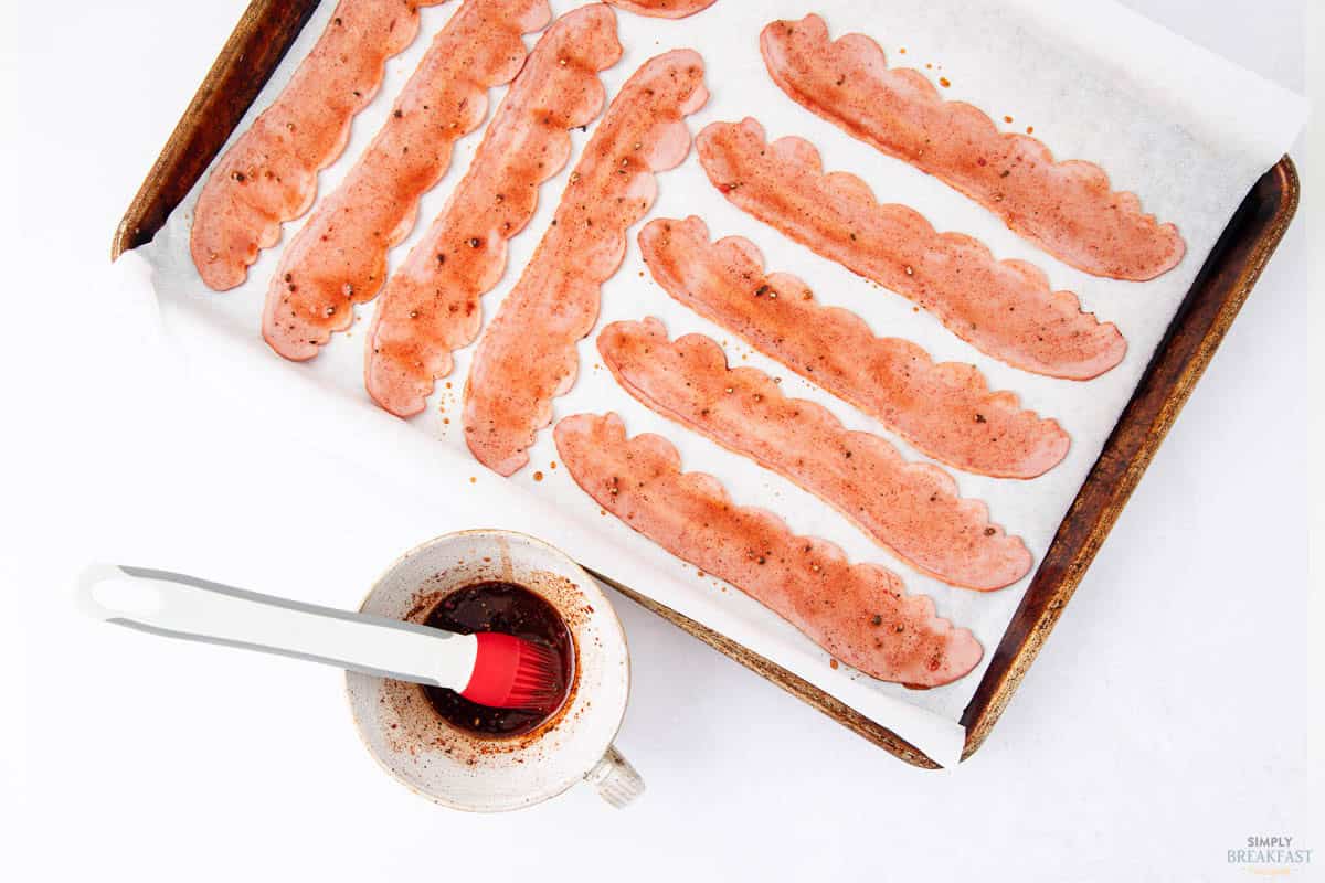 Slices of turkey bacon are arranged on a baking tray lined with parchment paper. A white bowl with a basting brush, coated with a reddish marinade, is placed beside the tray.