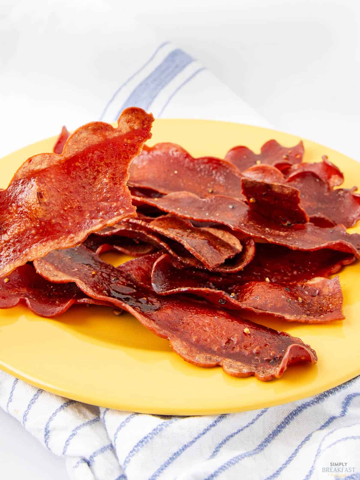 A yellow plate filled with crispy bacon strips sits on a blue and white striped cloth. The bacon appears golden and slightly curled, contrasting with the bright plate.