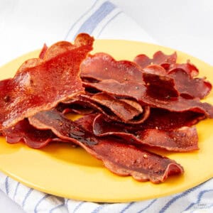 Crispy strips of vegan bacon made from carrot, arranged on a yellow plate placed on a white and blue striped cloth. The bacon is slightly shiny, with a reddish color and wavy edges.