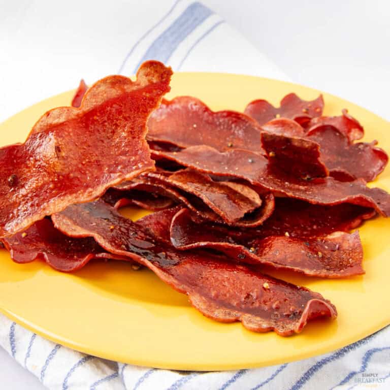 Crispy strips of vegan bacon made from carrot, arranged on a yellow plate placed on a white and blue striped cloth. The bacon is slightly shiny, with a reddish color and wavy edges.