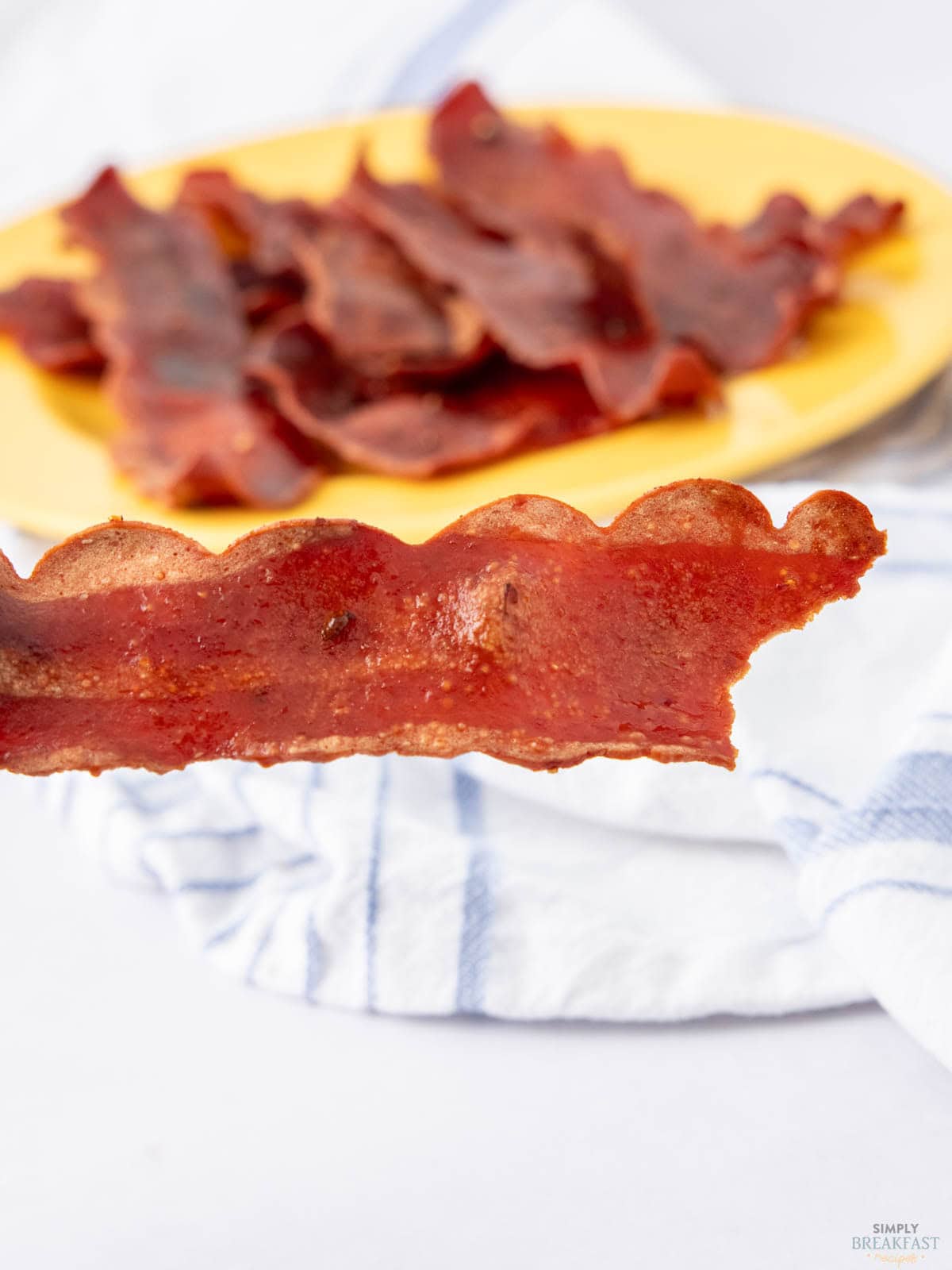 A close-up of a crispy piece of bacon held in focus in front of a blurred plate with more bacon. A white and blue striped cloth is partially visible in the background.