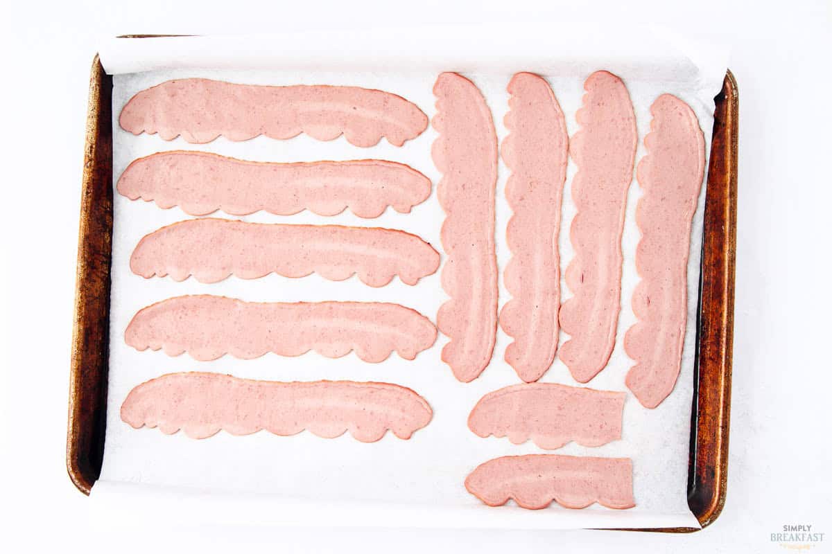 Strips of vegetarian bacon arranged on a parchment-lined baking sheet, ready for cooking. The strips have a pinkish hue and vary in length, placed neatly in rows.