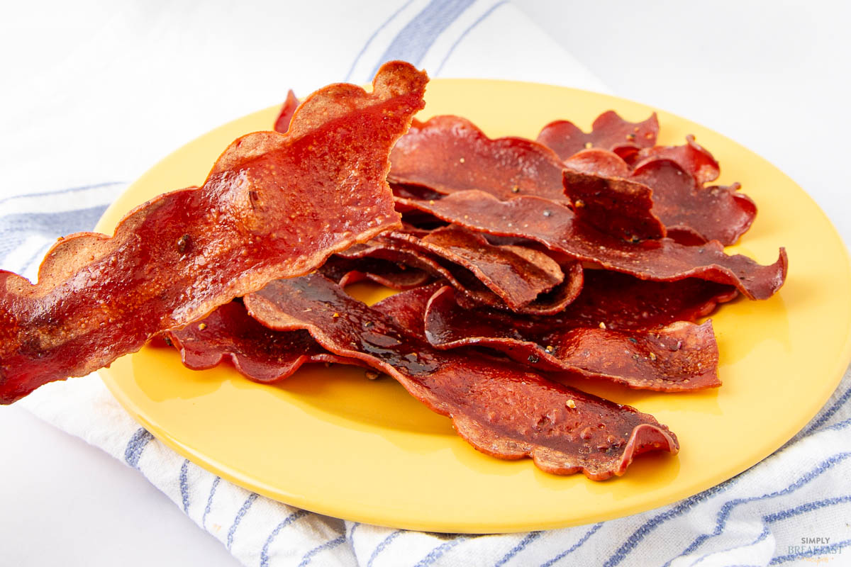 A plate of crispy, browned bacon strips is arranged on a yellow dish. One slice is held up, showing its wavy texture. A blue and white striped towel is partially visible underneath the plate.