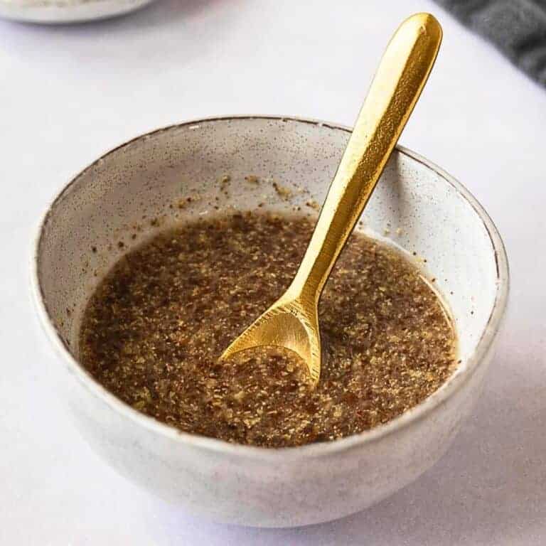 A white ceramic bowl containing a thick brown mixture, with a gold spoon resting inside. The background is a soft white surface, and a hint of another bowl is visible in the upper left corner.