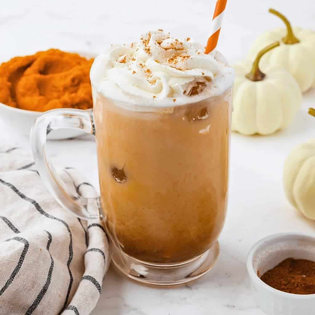 A glass mug of iced pumpkin spice latte topped with whipped cream and cinnamon, with a striped straw. Surrounding items include a bowl of pumpkin puree, small white pumpkins, cinnamon powder, and a striped cloth.