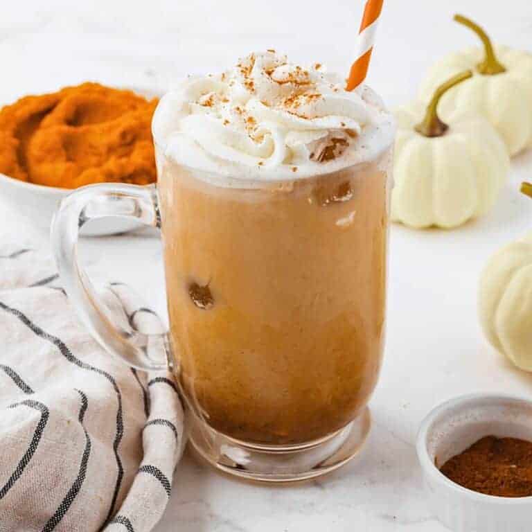 A glass mug of iced pumpkin spice latte topped with whipped cream and cinnamon, with a striped straw. Surrounding items include a bowl of pumpkin puree, small white pumpkins, cinnamon powder, and a striped cloth.