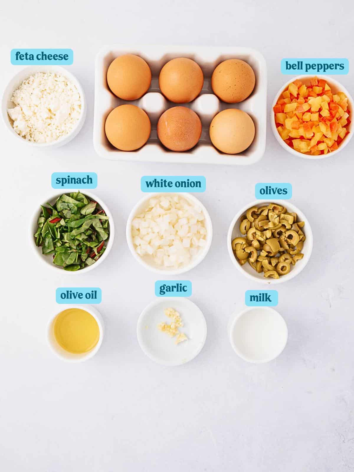 Top-down view of ingredients for a dish including feta cheese, eggs, diced bell peppers, chopped spinach, chopped white onion, sliced olives, olive oil, minced garlic, and milk arranged in bowls on a light surface.