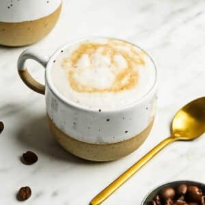 A speckled white and brown ceramic mug filled with frothy coffee topped with caramel swirl. A gold spoon lies beside it on a marble surface, with scattered coffee beans and a bowl of beans nearby.