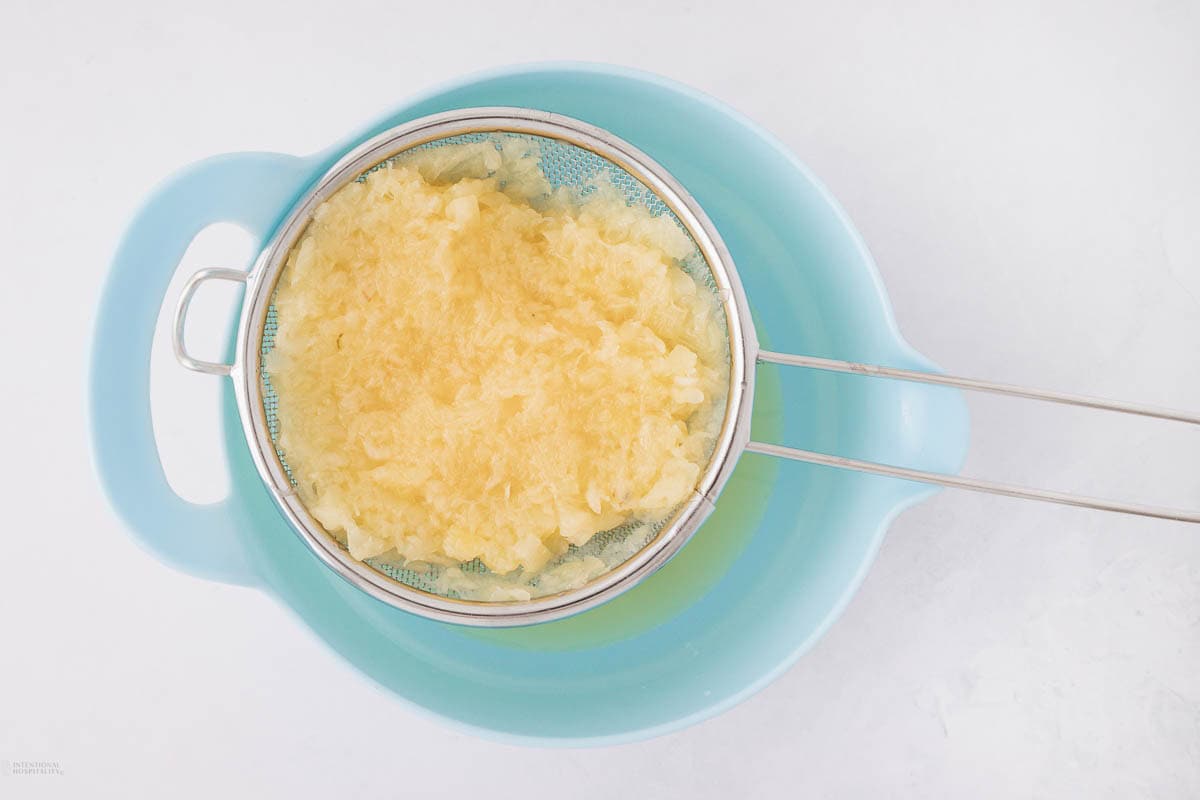 Finely grated pineapple being strained in a metal sieve over a blue bowl.