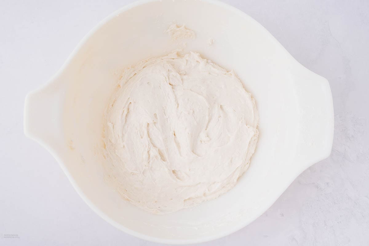 A white bowl filled with pale, smooth dough sits on a light-colored countertop. The dough appears freshly mixed and ready for rising or use in baking.