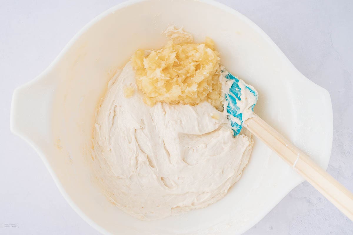 A white mixing bowl containing batter with a portion of crushed pineapple on top. A blue and white spatula rests on the edge of the bowl, partially submerged in the mixture. The background is a white surface.