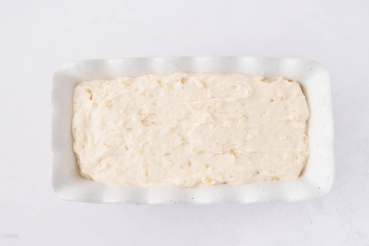 A rectangular baking dish filled with smooth, off-white batter, possibly for bread or cake, on a light background. The batter shows a creamy texture with small visible pieces scattered throughout.