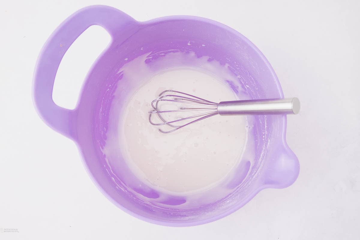 A purple mixing bowl containing creamy white batter with a metal whisk inside, set on a white surface.