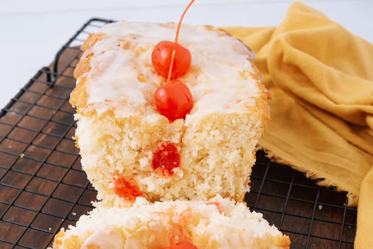 A glazed loaf cake with cherries on top and some inside is placed on a cooling rack. A slice is cut from the front, revealing the cake's soft texture. A yellow cloth is partially visible beside the cake.