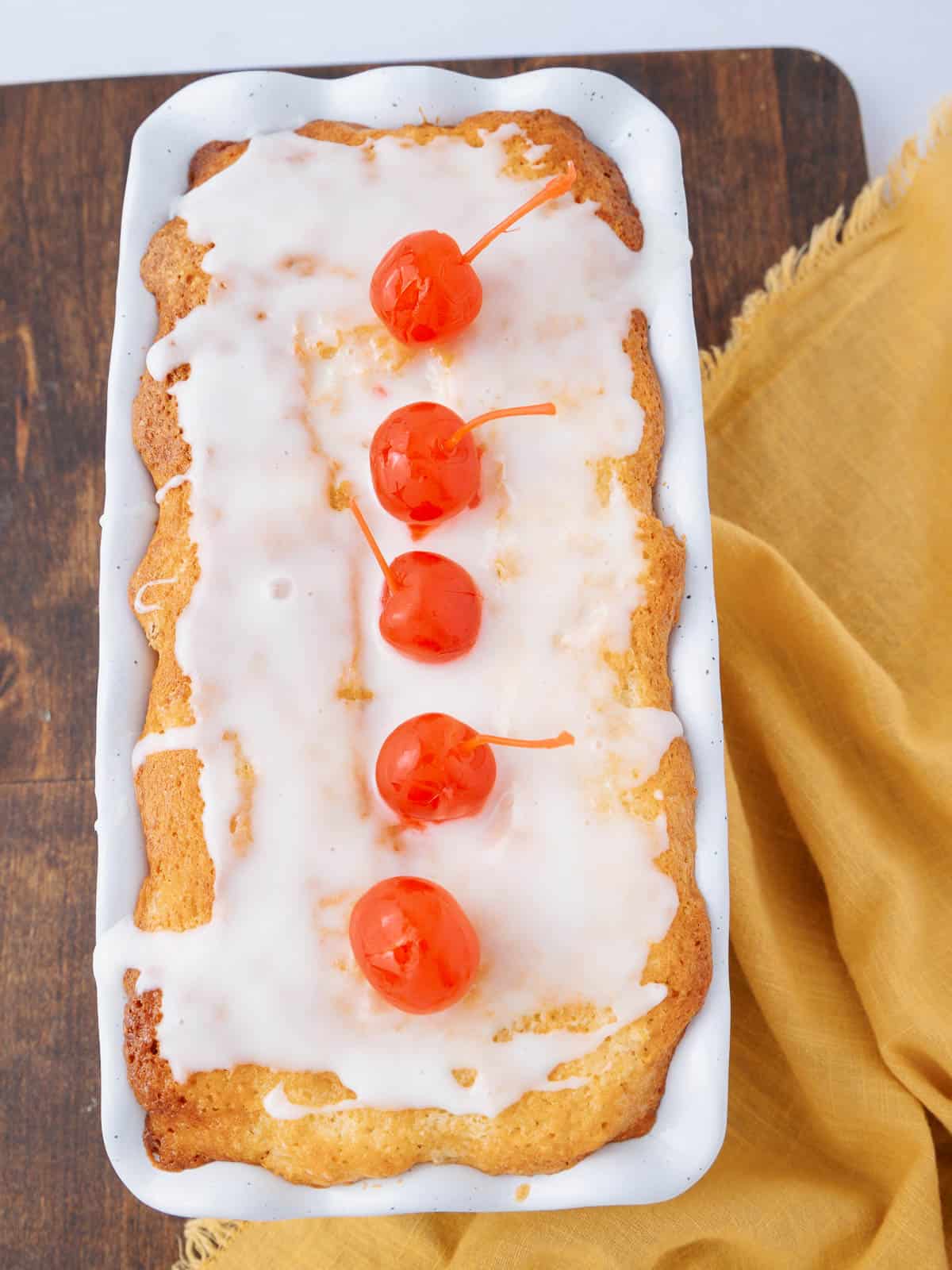 A loaf cake in a white dish drizzled with white icing and topped with five maraschino cherries in a row. The dish is on a wooden surface next to a yellow cloth.