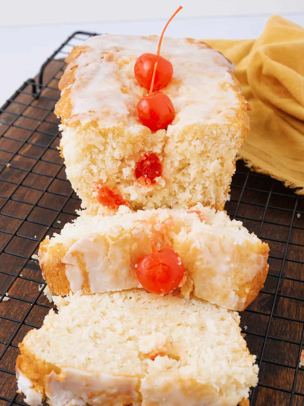 A loaf of glazed cherry bread on a cooling rack. The bread is topped with three bright red cherries and sliced to reveal a moist texture with cherries inside. A yellow cloth is partially visible in the background.