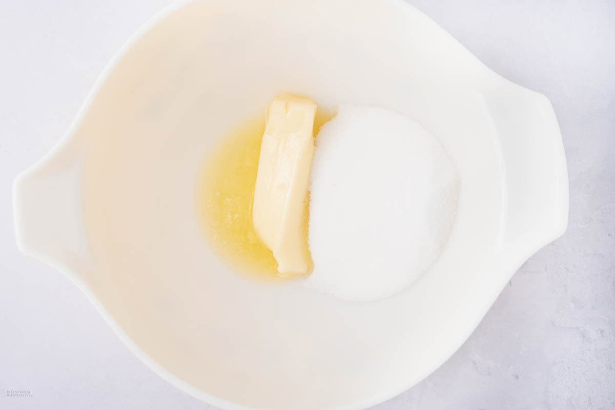 A white mixing bowl containing a stick of butter and granulated sugar, placed on a light surface.