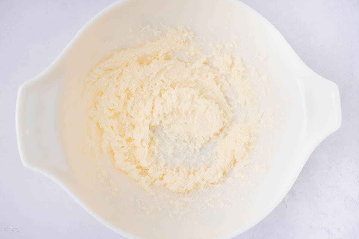 A white bowl contains a creamy mixture of butter and sugar, with a light and fluffy texture, against a light background.