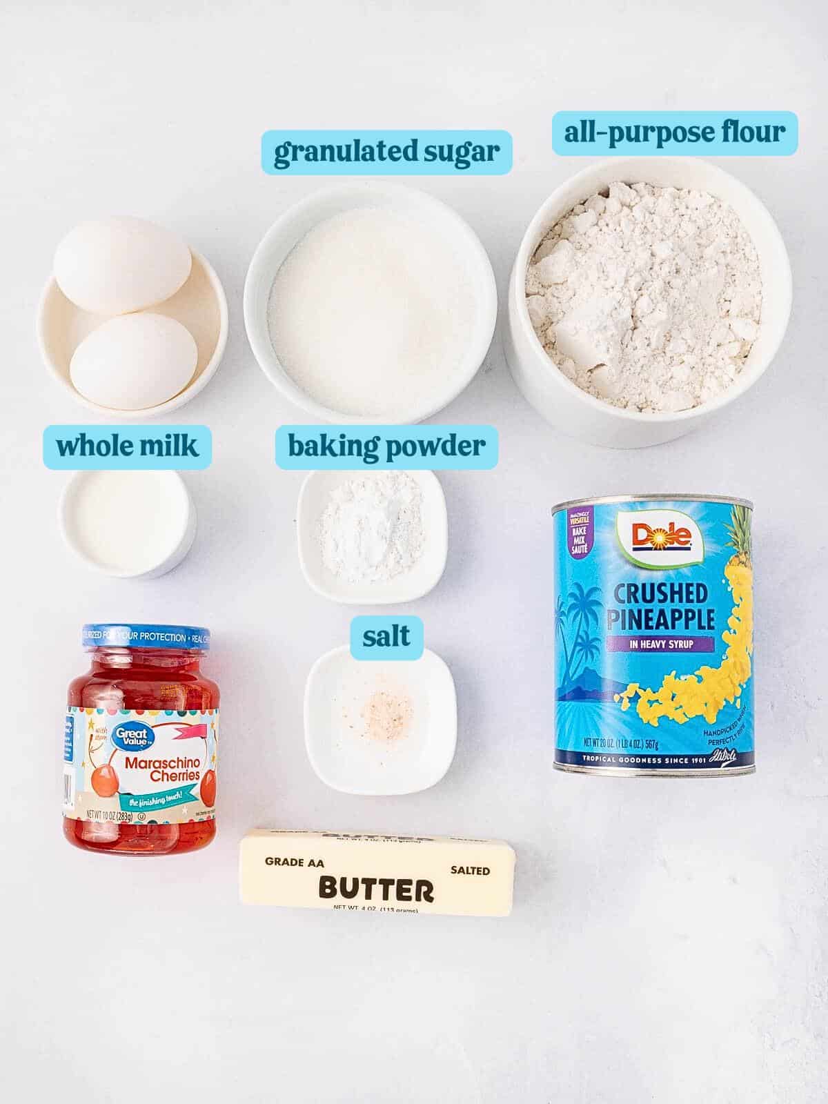 Ingredients for a recipe laid out: three eggs, whole milk, baking powder, granulated sugar, all-purpose flour, salt, butter, a jar of maraschino cherries, and a can of crushed pineapple.