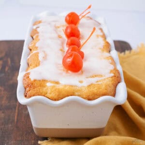 Loaf cake with white icing and four red cherries on top, placed in a decorative white loaf pan. The pan is on a wooden surface with a mustard-colored cloth beside it.