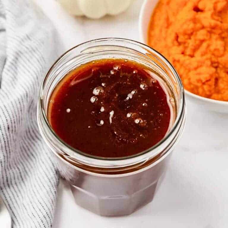 A jar filled with dark brown pumpkin spice syrup, placed in front of an orange pumpkin puree in a bowl. A striped cloth is beside them on a white surface.