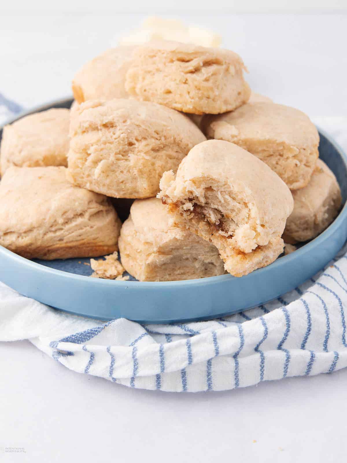 A blue plate filled with freshly baked biscuits stacked on top of a blue and white striped cloth. One biscuit has a bite taken out, revealing a fluffy inside.