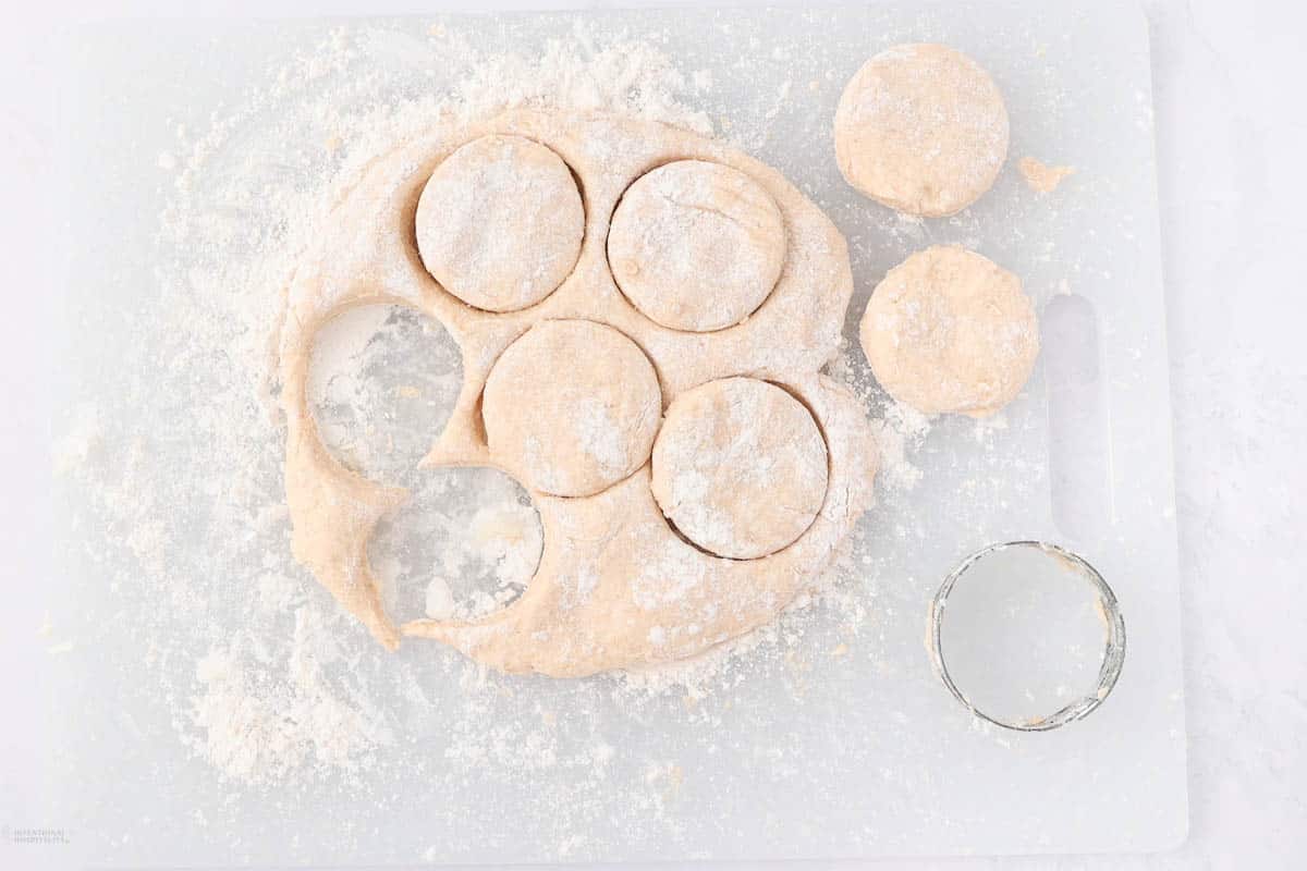 Dough on a cutting board with circular shapes cut out, dusted with flour. A metal cutter is seen nearby, along with leftover dough scraps.