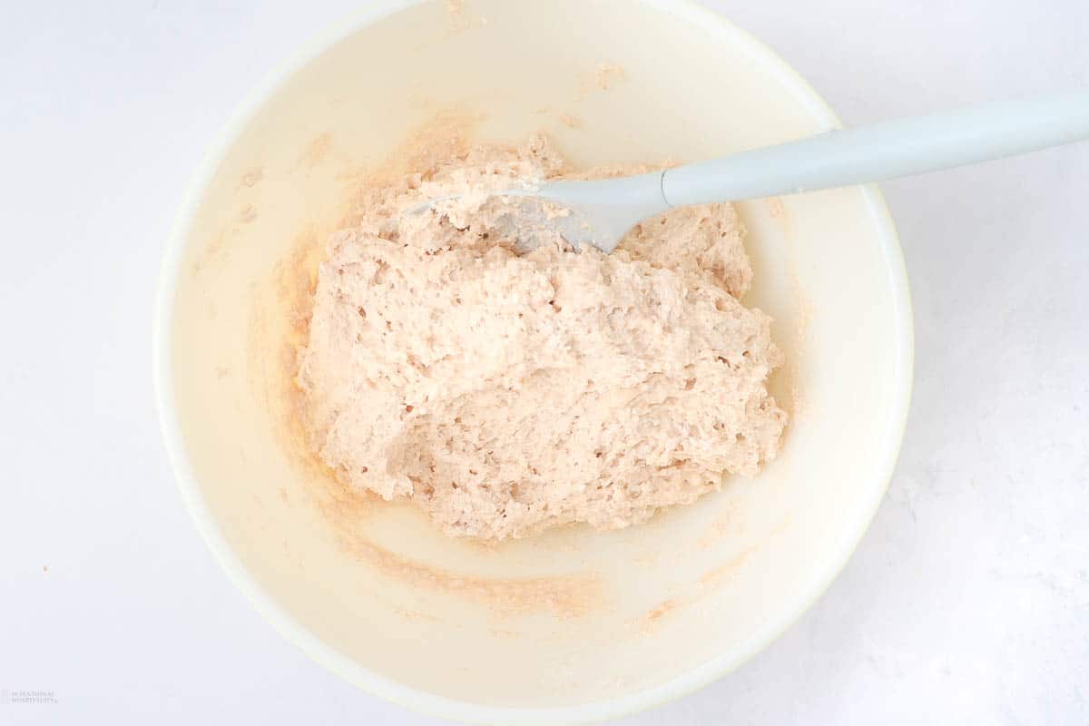 A bowl of raw bread dough is being mixed with a light blue spatula. The dough appears soft and unbaked, set in a creamy white bowl on a white surface.