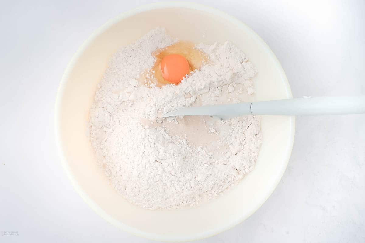 A white mixing bowl containing flour and a cracked egg. A white spatula rests in the bowl. The background is a light surface.