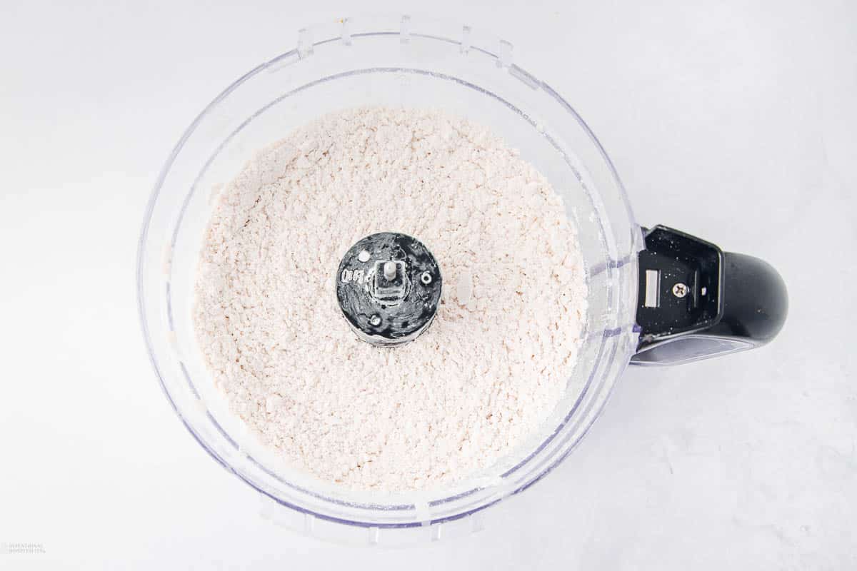 Top view of a food processor containing finely ground white flour, set against a light gray background. The black center blade is visible, surrounded by the even texture of the flour.