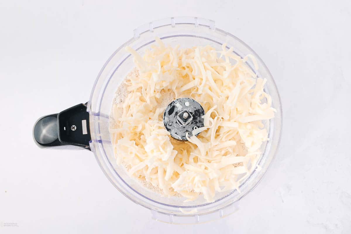 A transparent food processor bowl filled with shredded cheese, with a central black blade, placed on a white surface.