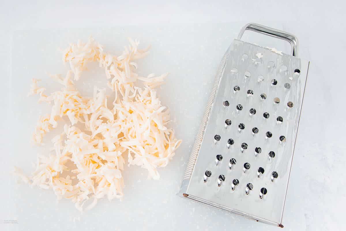 A metal box grater lies on a white surface next to a small pile of shredded white cheese.