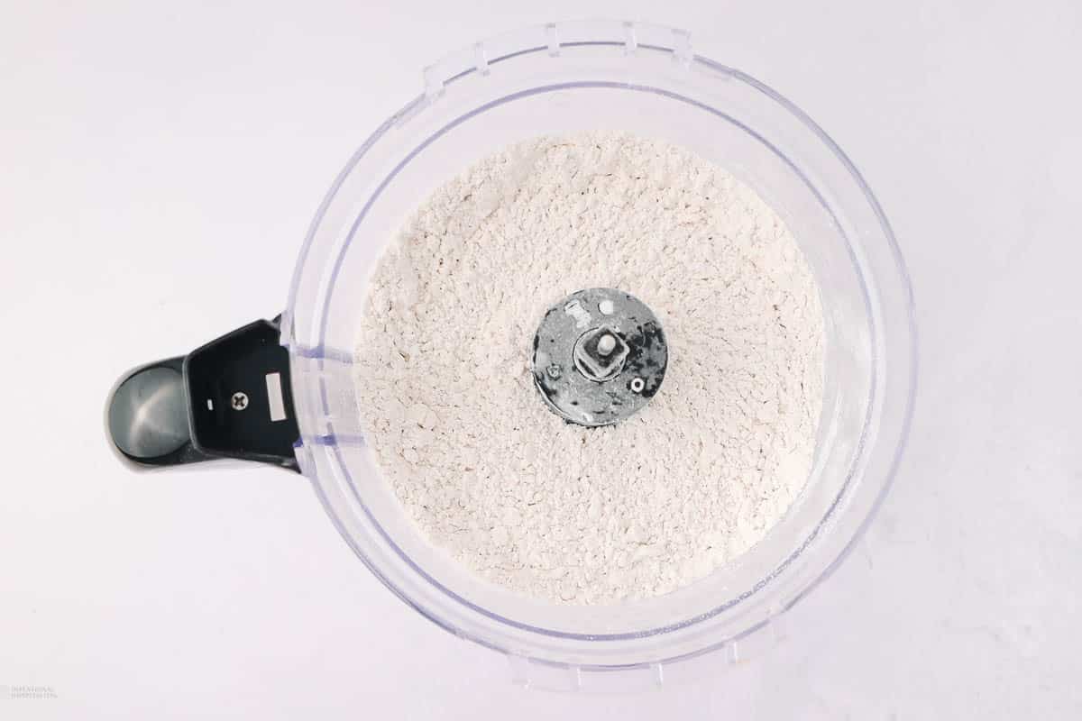 A top-down view of a food processor containing finely ground flour. The appliance is placed on a white surface, and the blades are visible in the center of the processed flour.