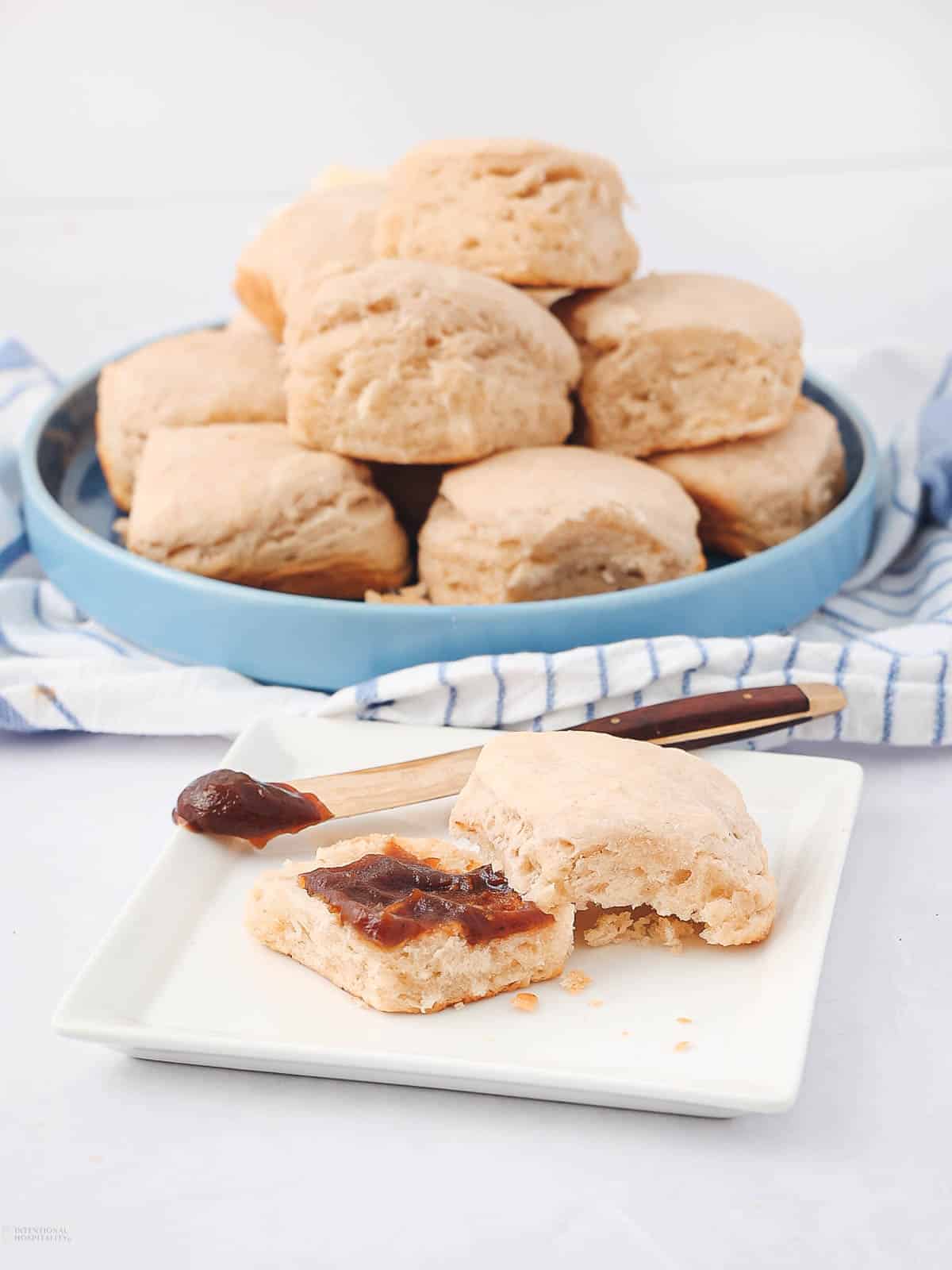 A plate of biscuits stacked high on a blue dish, with one biscuit on a square white plate in the foreground. The biscuit is partially eaten, topped with jam, accompanied by a knife with more jam on the blade. A striped cloth is underneath.