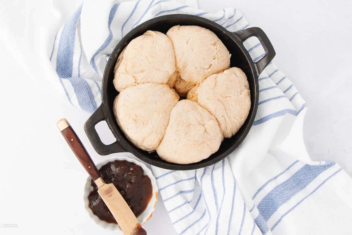 A skillet with six uncooked biscuit dough pieces, placed on a white and blue striped cloth. Beside it, there's a small bowl of dark spread with a knife resting on the edge.