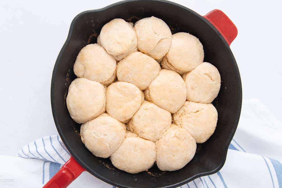 A cast iron skillet filled with unbaked biscuit dough arranged in a neat pattern. The skillet has a red handle and rests on a blue and white striped cloth on a white surface.