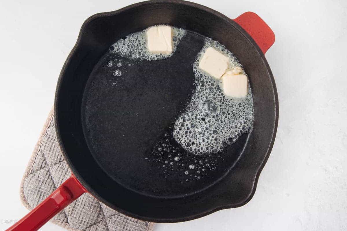 A black skillet with a red handle on a beige checkered pot holder contains three melting cubes of butter, creating a foamy texture. The background is a clean white surface.