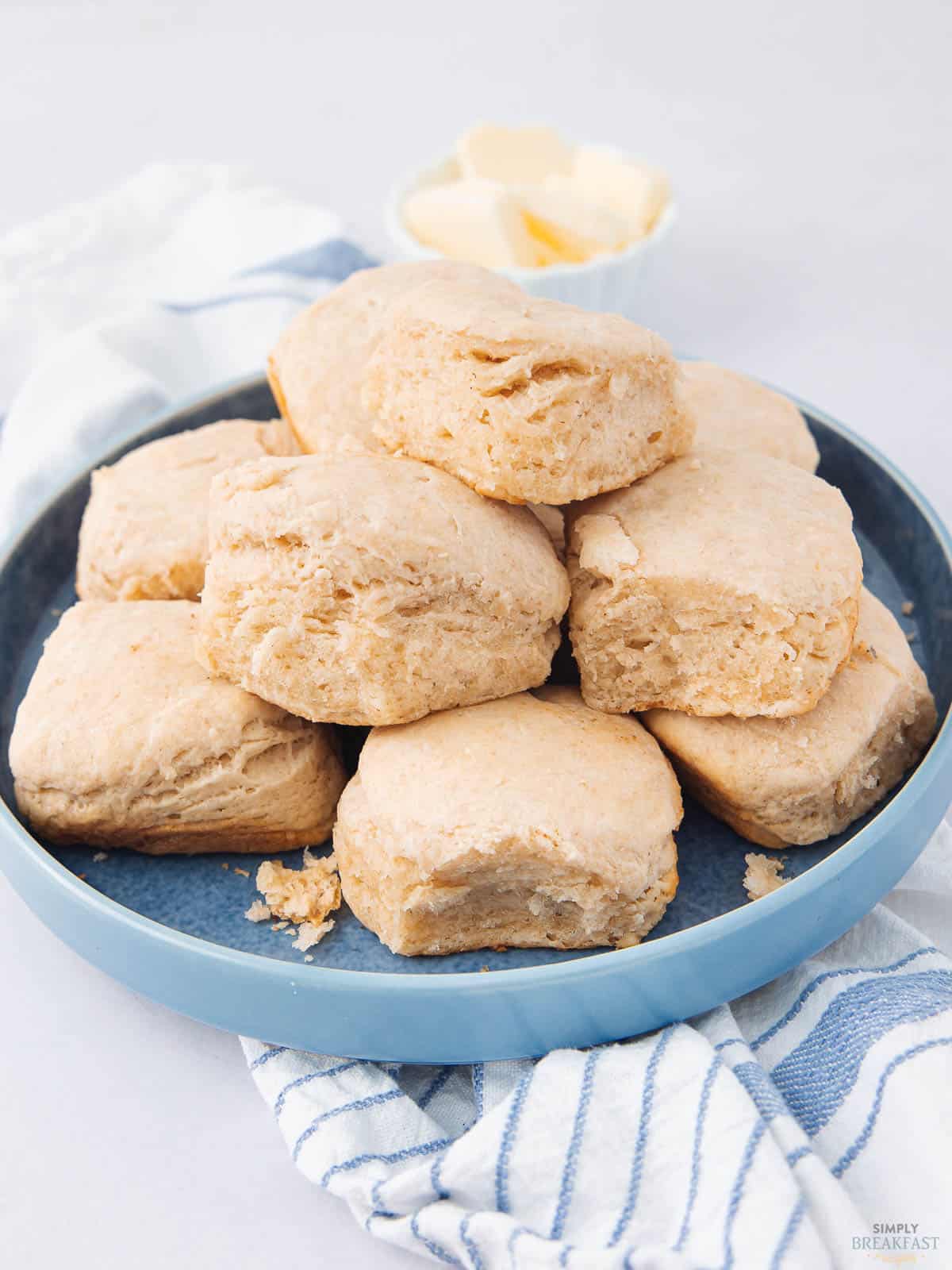 A blue plate stacked with fluffy, golden biscuits on a white surface. A blue and white striped cloth is partially under the plate. In the background, a small bowl contains butter pieces.