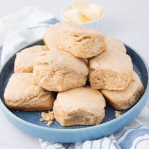 A blue plate piled with freshly baked, flaky biscuits. A small dish with butter is in the background, and a striped cloth rests beneath the plate on a light surface.