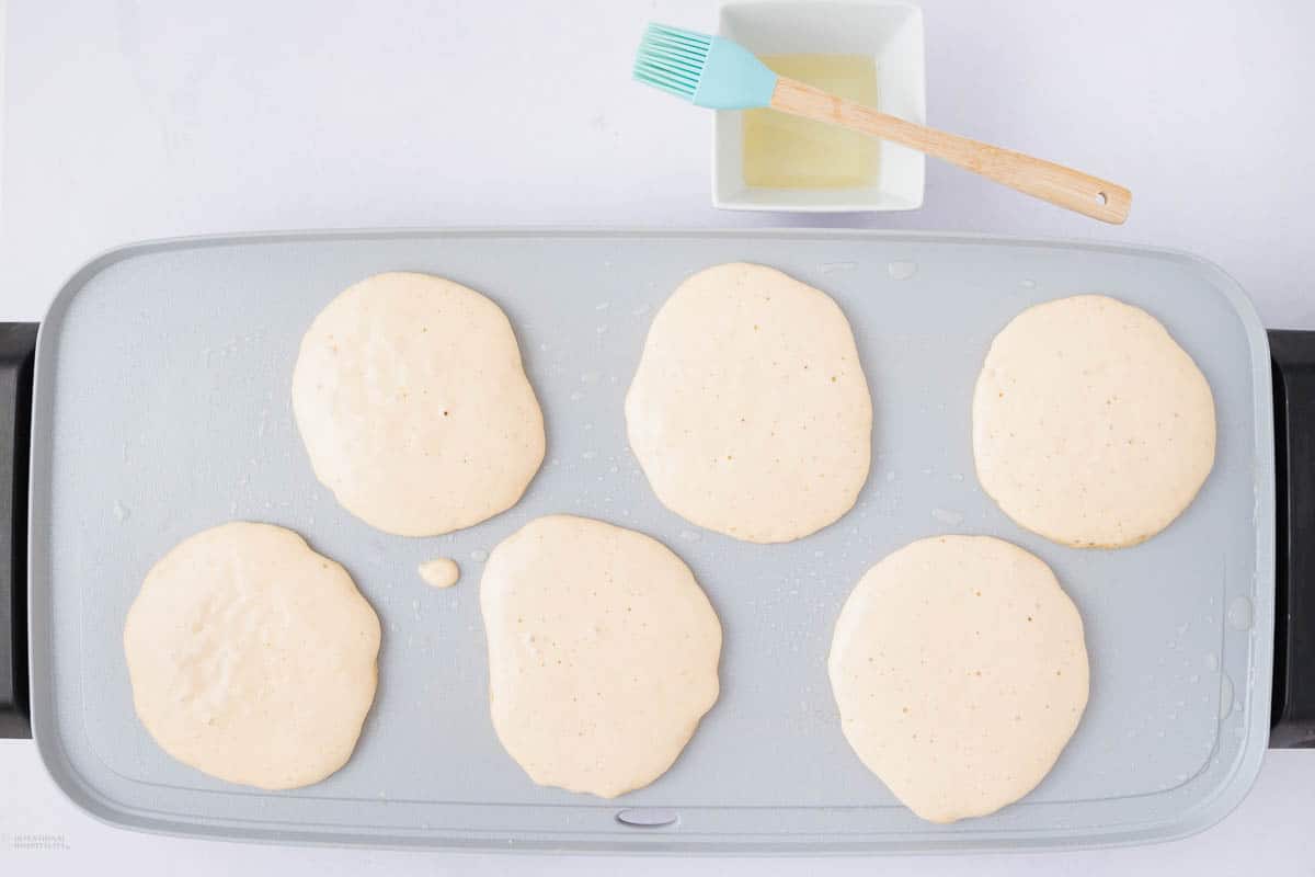 A flat stovetop griddle with six circles of pancake batter cooking. A small brush and a white square bowl with oil or butter are placed above the griddle.