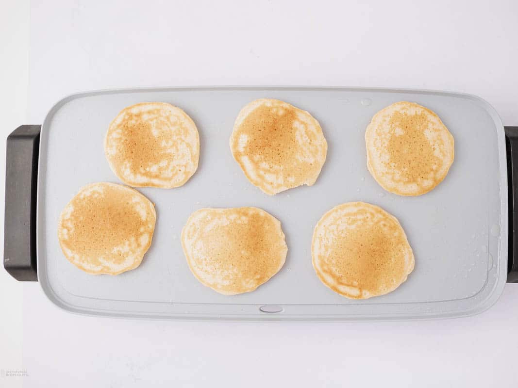 Six golden pancakes are cooking on a rectangular griddle.