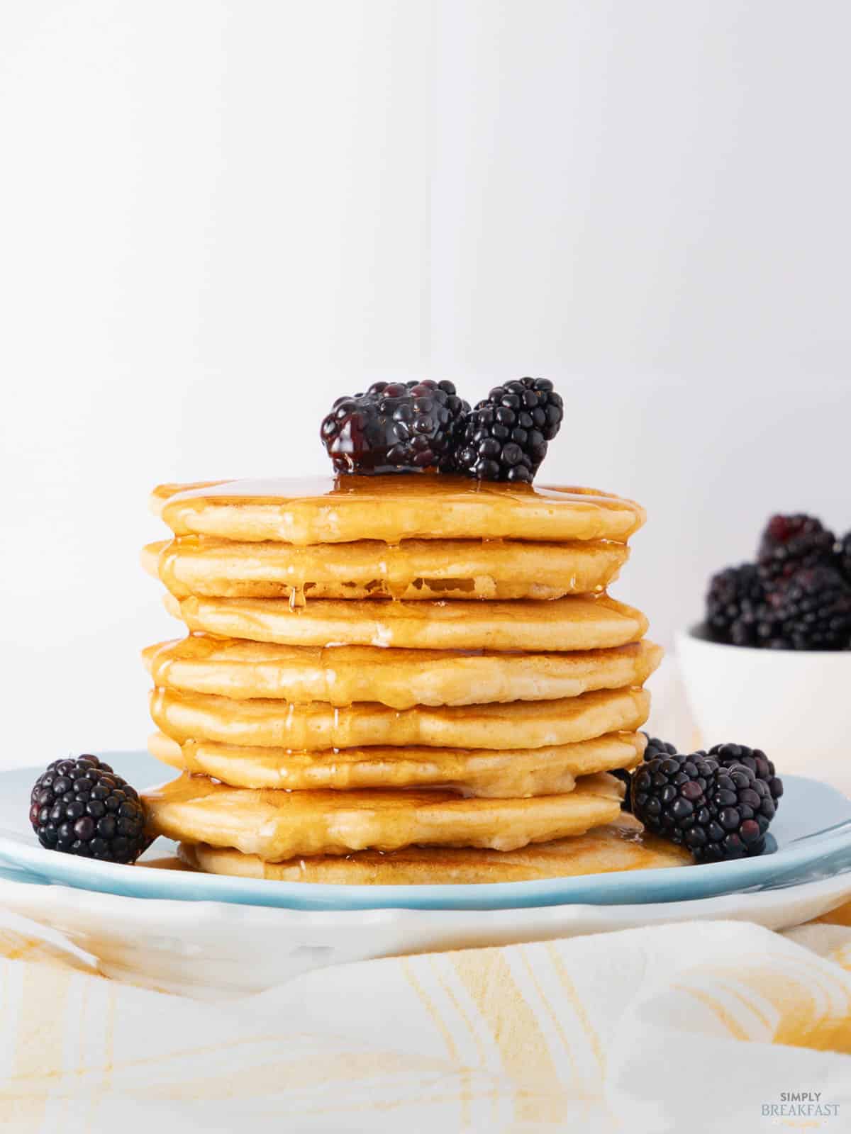 A stack of fluffy pancakes topped with fresh blackberries and drizzled with syrup, served on a blue plate. A bowl of blackberries is in the background, with a striped yellow and white cloth beneath the plate.