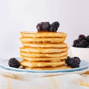 A stack of golden pancakes drizzled with syrup, topped with blackberries on a white plate. A bowl of blackberries is in the background on a light surface.